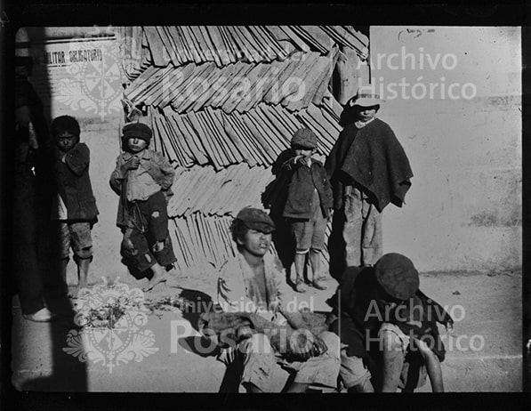 Seis niños frente a unas filas acumuladas de tejas de barro, con las que se cubrían los techos de las casas más sólidas de las ciudades. Los niños aparecen con ropas desgastadas, descalzos; otros con ruanas y boinas. En el cartel de la pared, se alcanza a leer: "MILITAR OBLIGATORIO", probablemente haciendo referencia al servicio militar obligatorio. En las primeras décadas del siglo XX, el tema del servicio militar obligatorio fue objeto de debate en el país. En 1912, se expidió la ley que hacía obligatoria