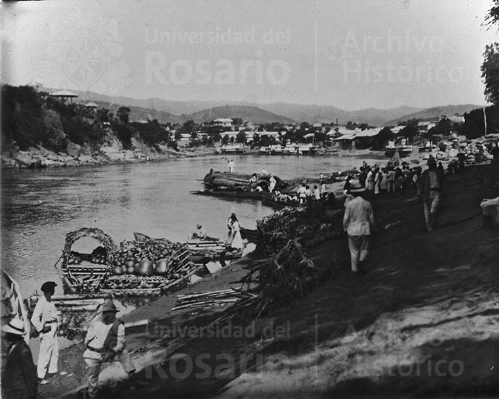 Puerto de Girardot, llamado De la Plaza, un lugar de afluencia del comercio de víveres. Se aprecia la ribera del río, colinas, embarcaciones pequeñas cargadas y varias personas esperando en la ribera. Al fondo, se ven algunas casas pequeñas alrededor del río.